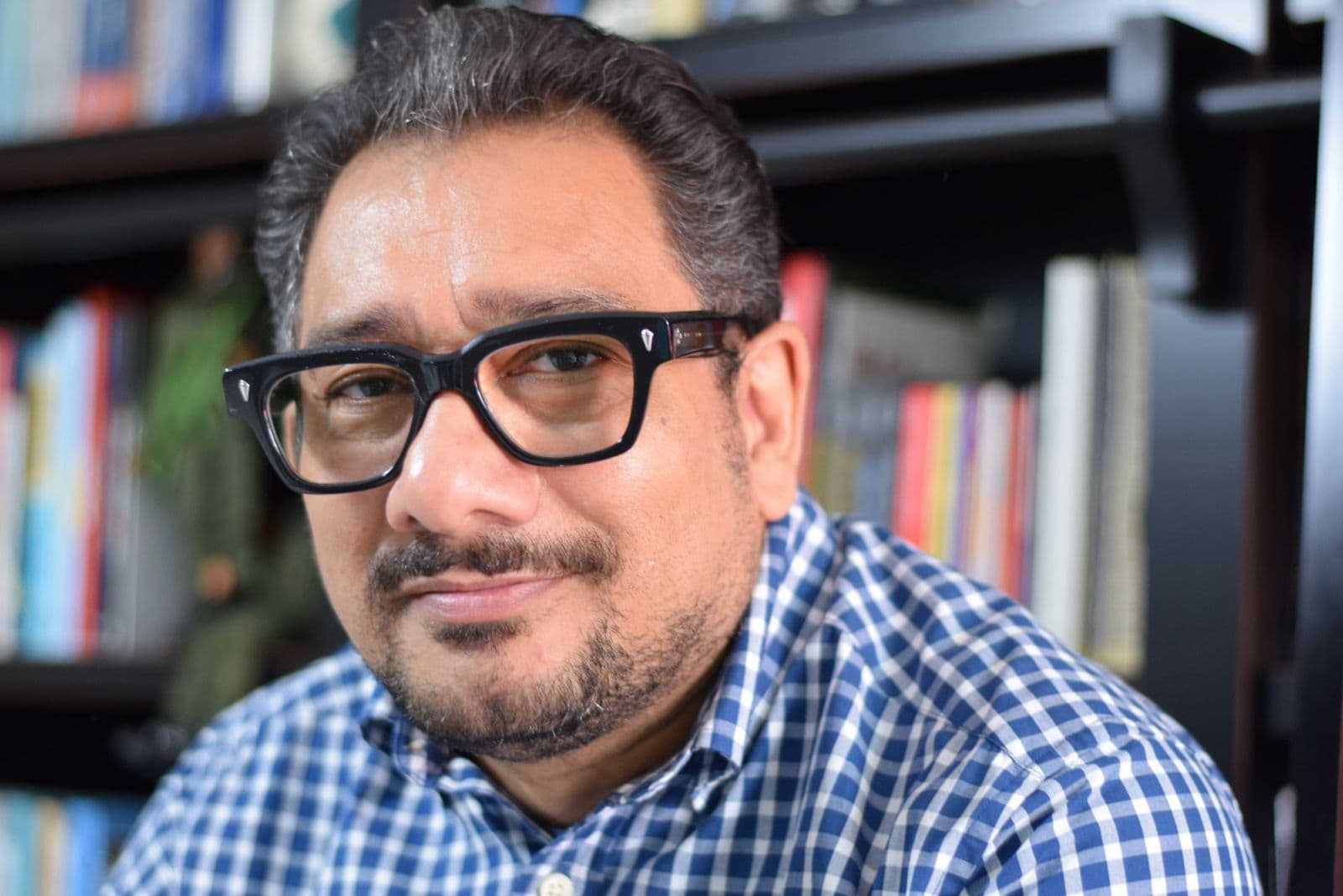Enzo Borges em retrato aproximado, de camisa xadrez, diante de uma estante de livros